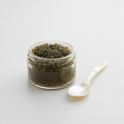 Close-up of fresh, high-quality caviar in a small glass jar, with a mother-of-pearl spoon beside it on a clean white background
